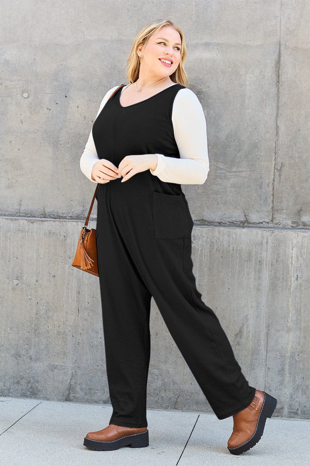 Woman wearing a black sleeveless straight jumpsuit with pockets, paired with a white shirt and brown shoes, carrying a handbag.