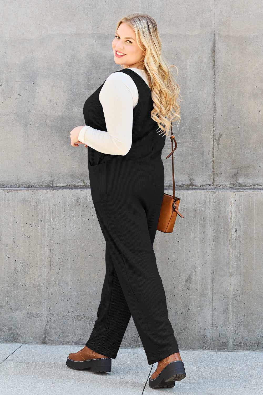 Woman wearing a sleeveless straight jumpsuit with pockets, styled with a brown crossbody bag and boots, made from polyester and spandex.