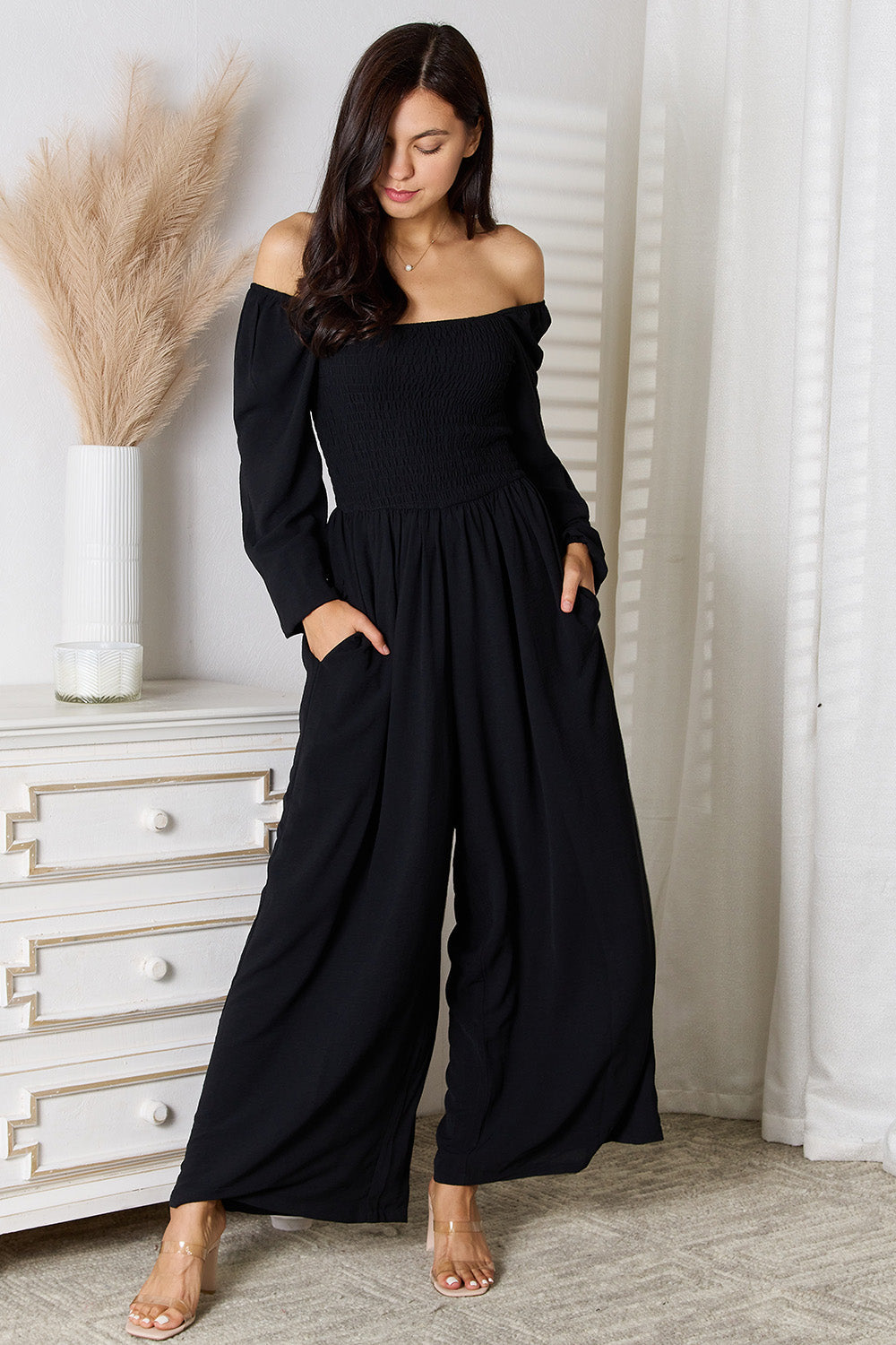 Woman wearing black square neck jumpsuit with pockets and smocked sheer bodice, standing in stylish room setting.