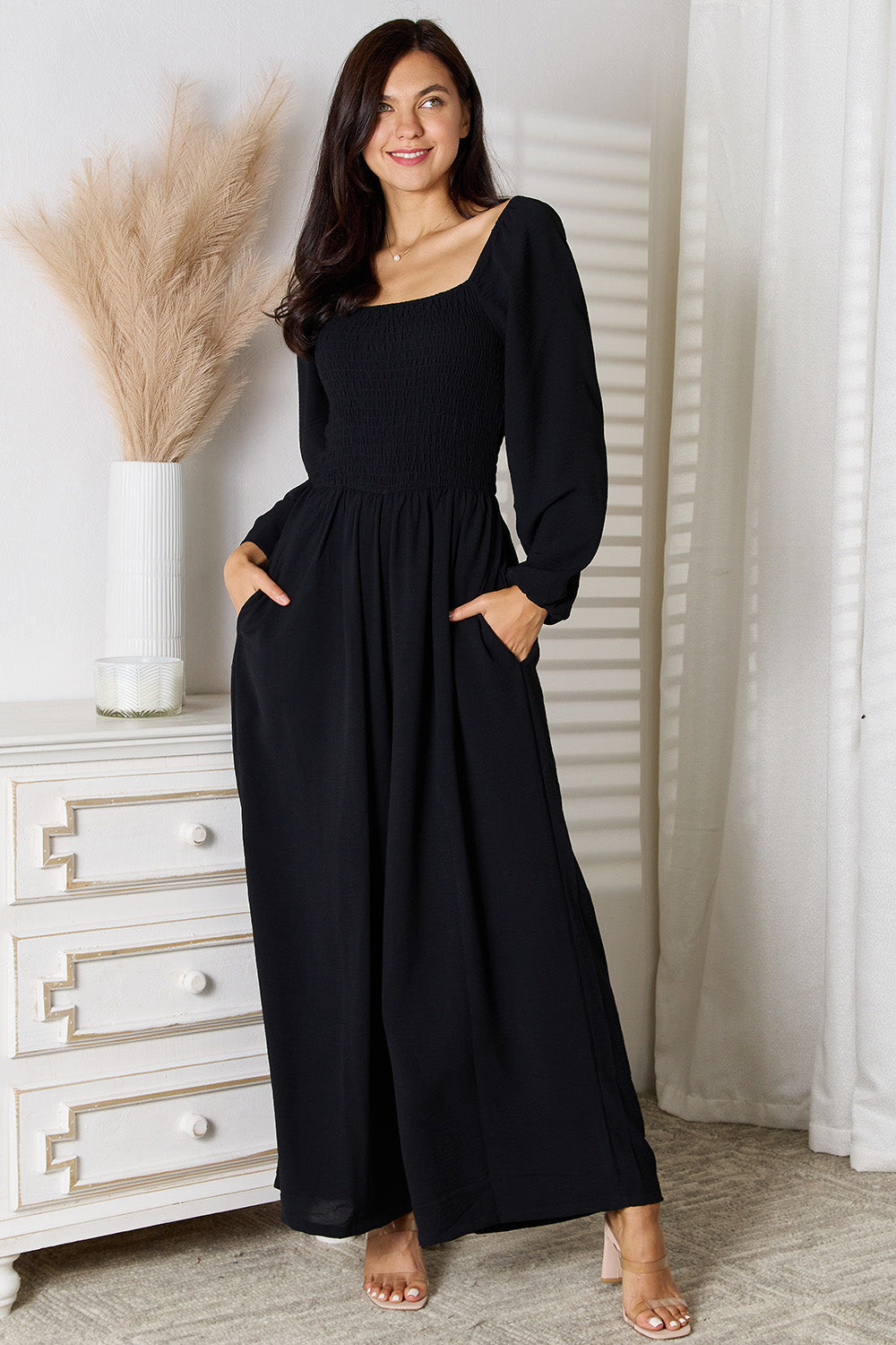 Woman wearing black square neck jumpsuit with pockets, smocked bodice, standing in a stylish room setting.