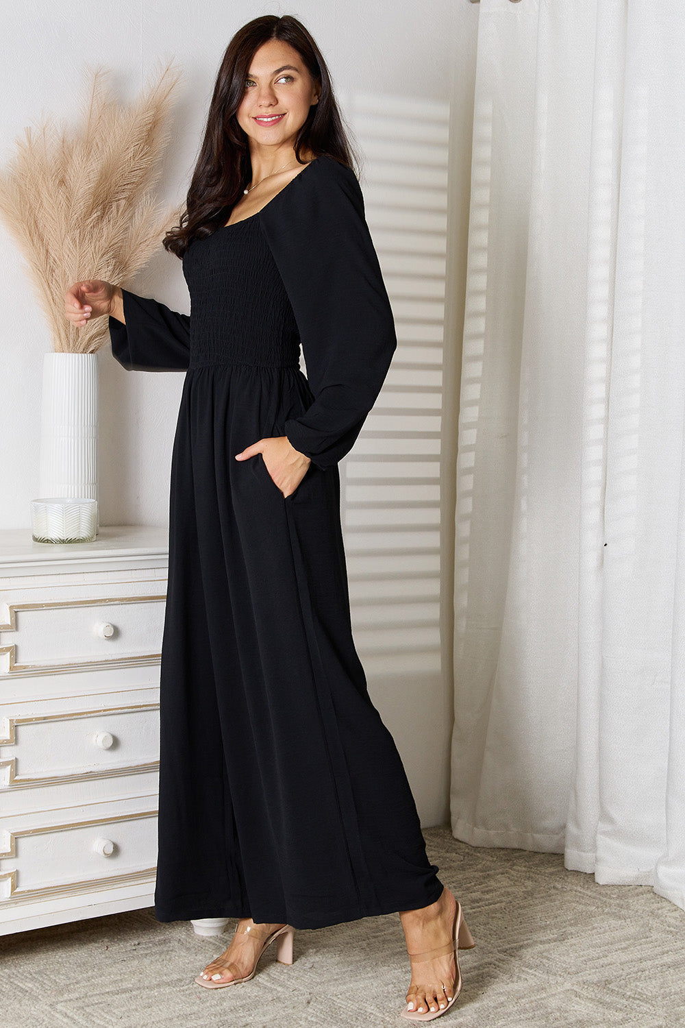 Woman wearing a black square neck jumpsuit with pockets, featuring a smocked sheer bodice, standing in a chic room.