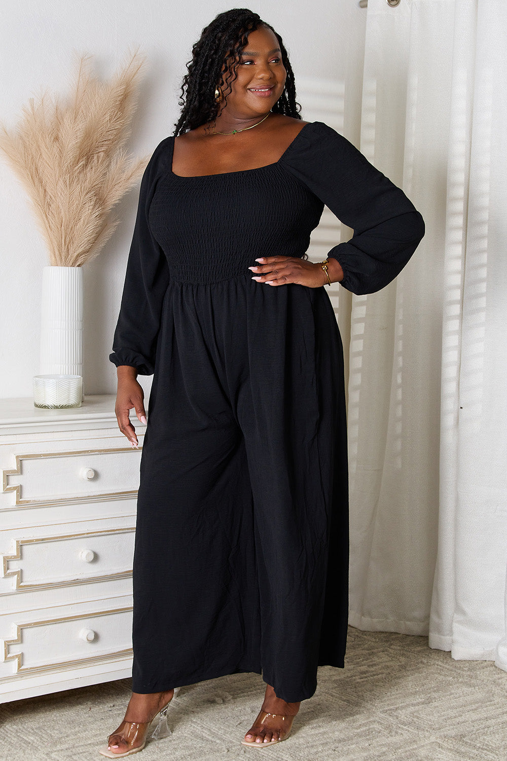 Woman wearing a black square neck jumpsuit with pockets, featuring a smocked sheer bodice, styled in a chic home setting.