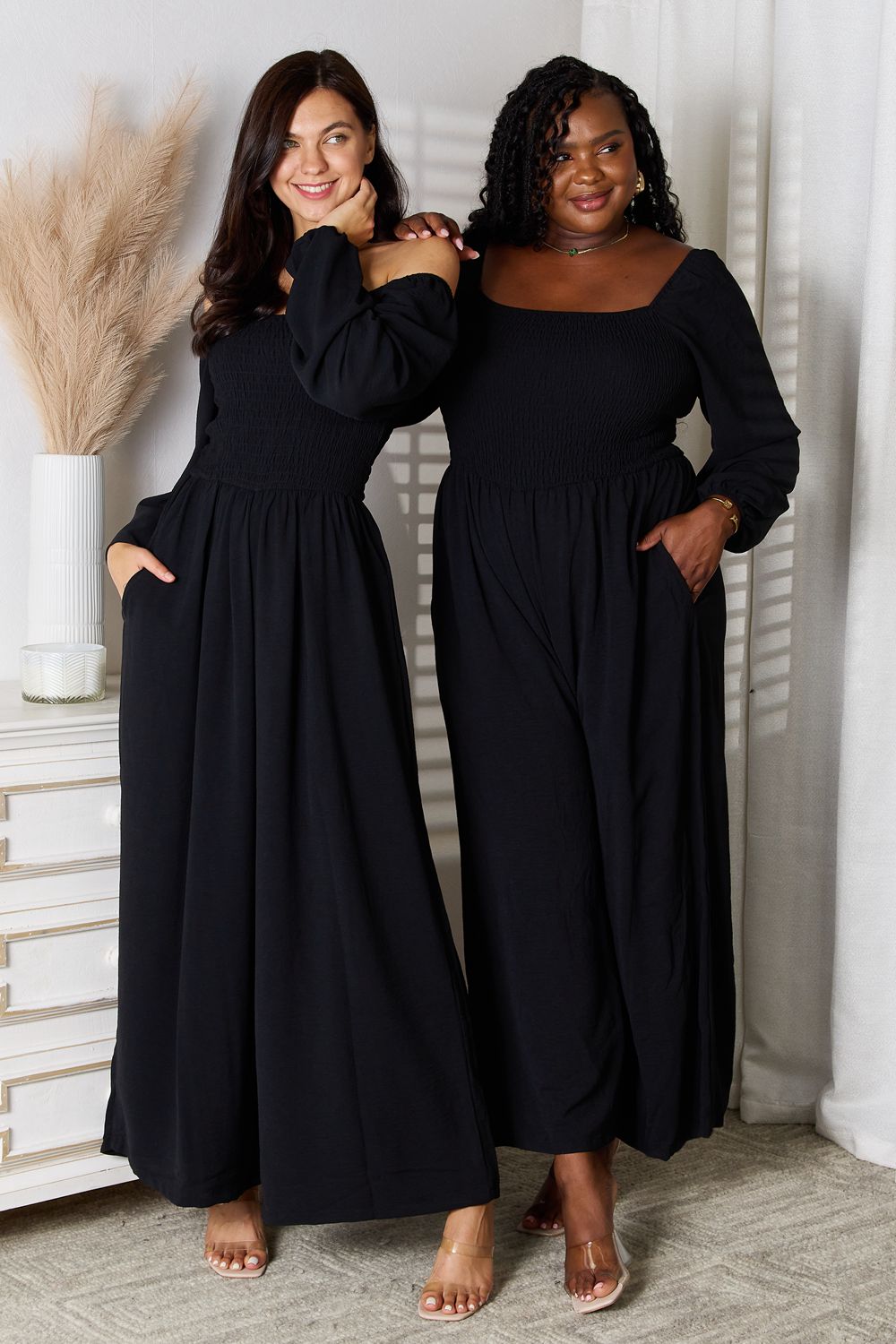 Two models wearing black square neck jumpsuits with pockets and smocked sheer bodice, showcasing flattering fit for all sizes.