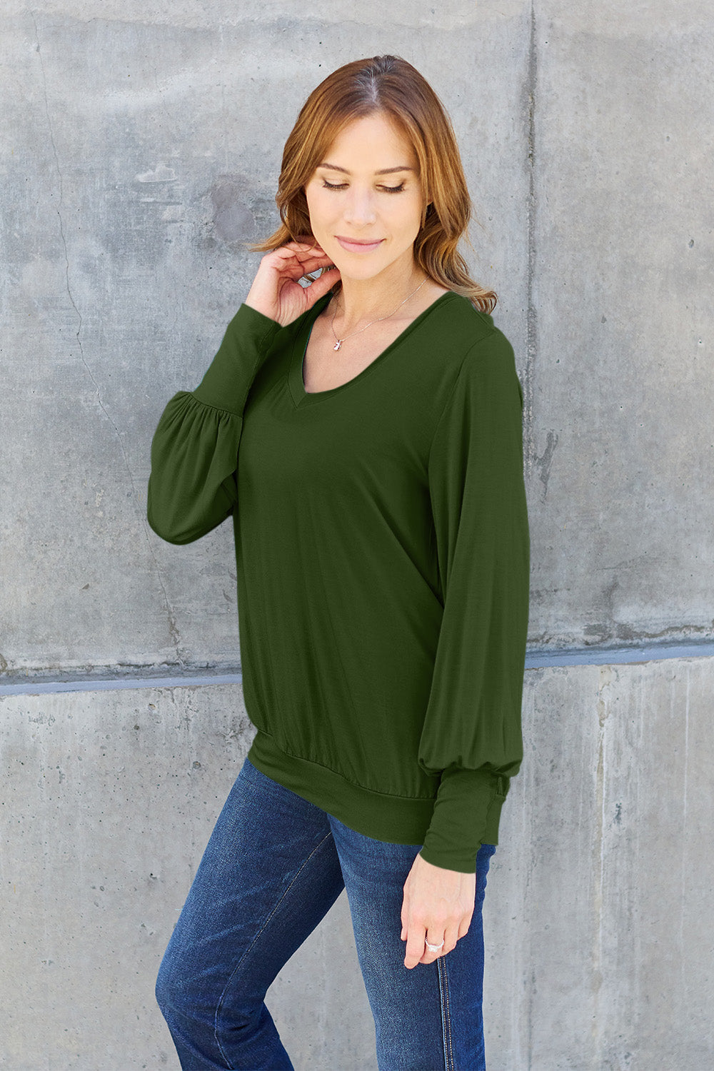 Woman wearing a green V-neck lantern sleeve top made of rayon and spandex, paired with jeans, standing against a concrete wall.