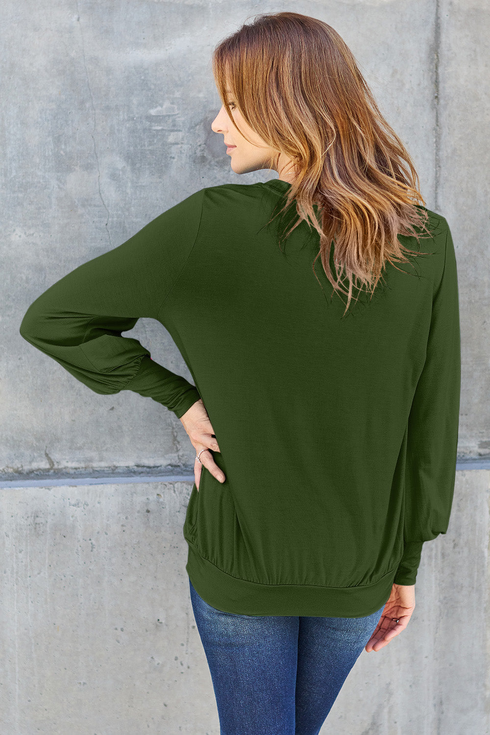 Woman wearing a green V-neck lantern sleeve top with slightly stretchy rayon blend, back view against concrete wall.