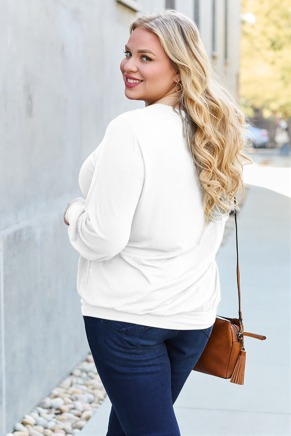 Woman wearing a white jacket with jeans holding a brown purse, smiling while walking down the street.