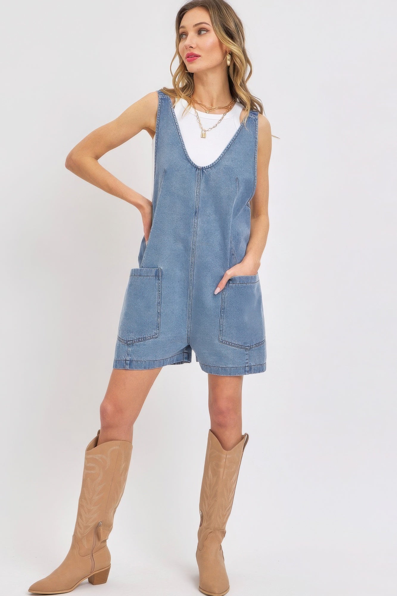 Woman wearing summer denim overall romper with oversized pockets and adjustable straps, styled with a white top and tan boots.