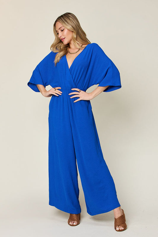 Blue surplice wide leg jumpsuit with pockets, featuring tied waistband and ruched details, made of polyester and spandex.