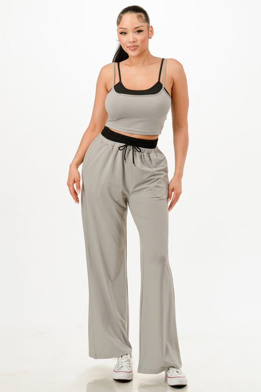 Woman in sage green crop top and matching wide-leg pants with black contrast trim, wearing white sneakers, standing against white background.