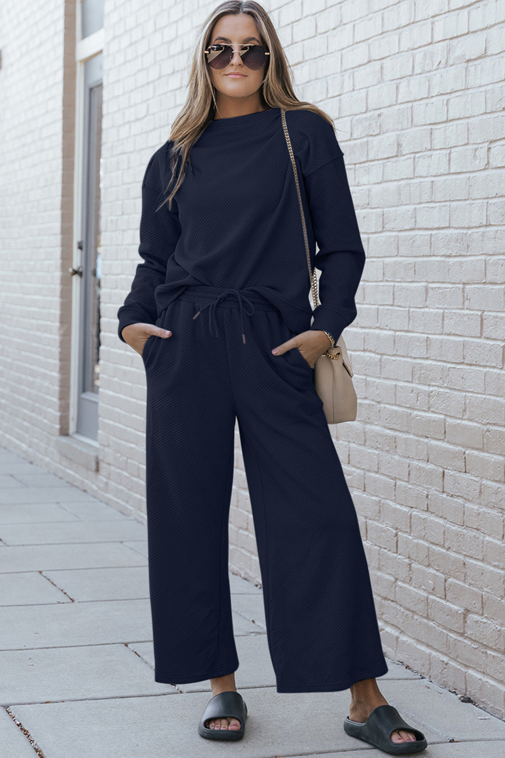 Woman wearing textured long sleeve top and drawstring pants set, accessorized with sunglasses and a beige shoulder bag, standing outside.