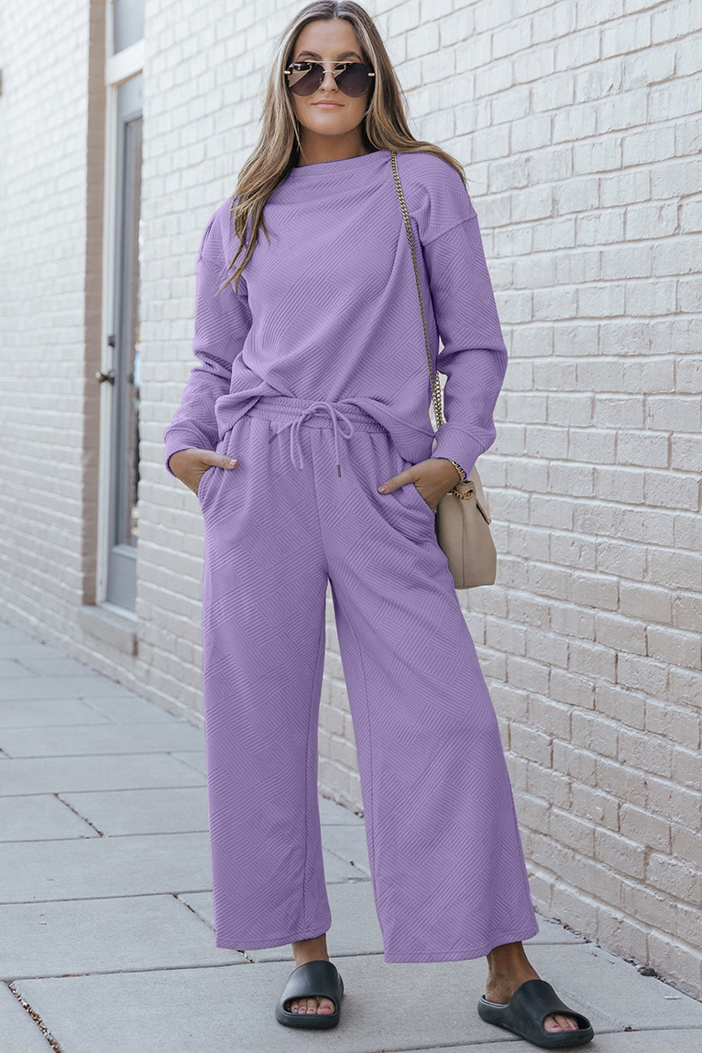 Woman wearing a textured long sleeve top and drawstring pants set in purple, standing outdoors with sunglasses and a handbag.