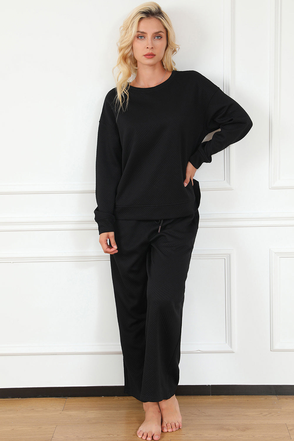 Woman in black textured long sleeve top and drawstring pants set, slightly stretchy fabric, standing against white wall.