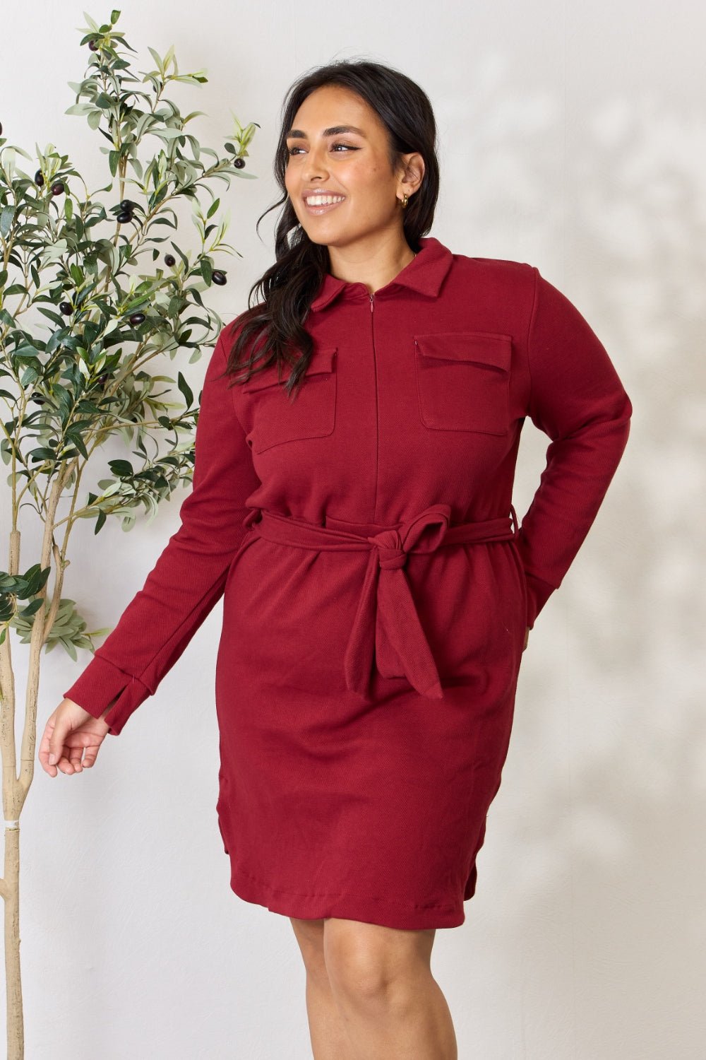 Woman wearing a red tie front half zip long sleeve shirt dress with pockets, standing near a plant.