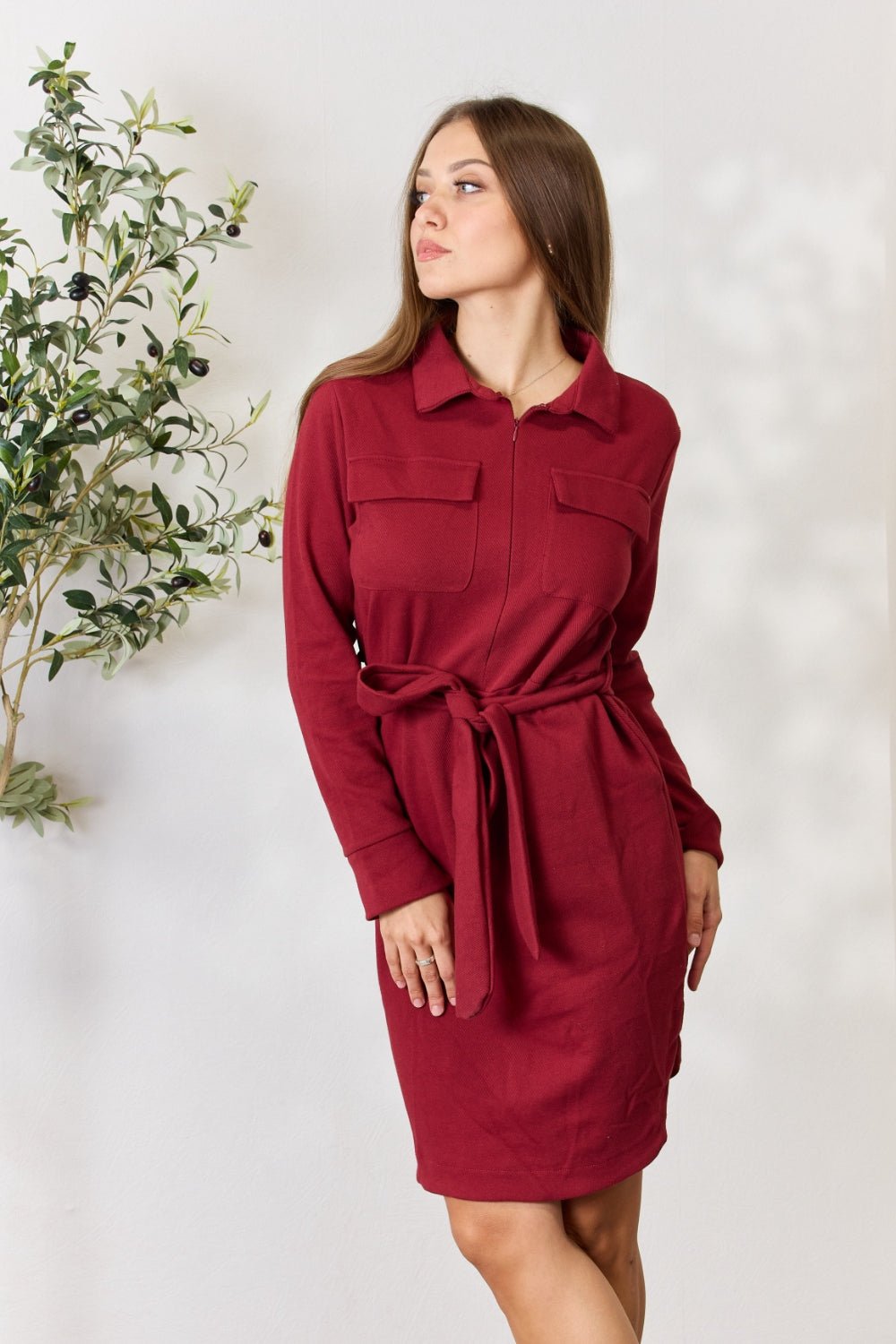 Woman wearing a maroon tie front half zip long sleeve shirt dress with pockets, standing near a plant.