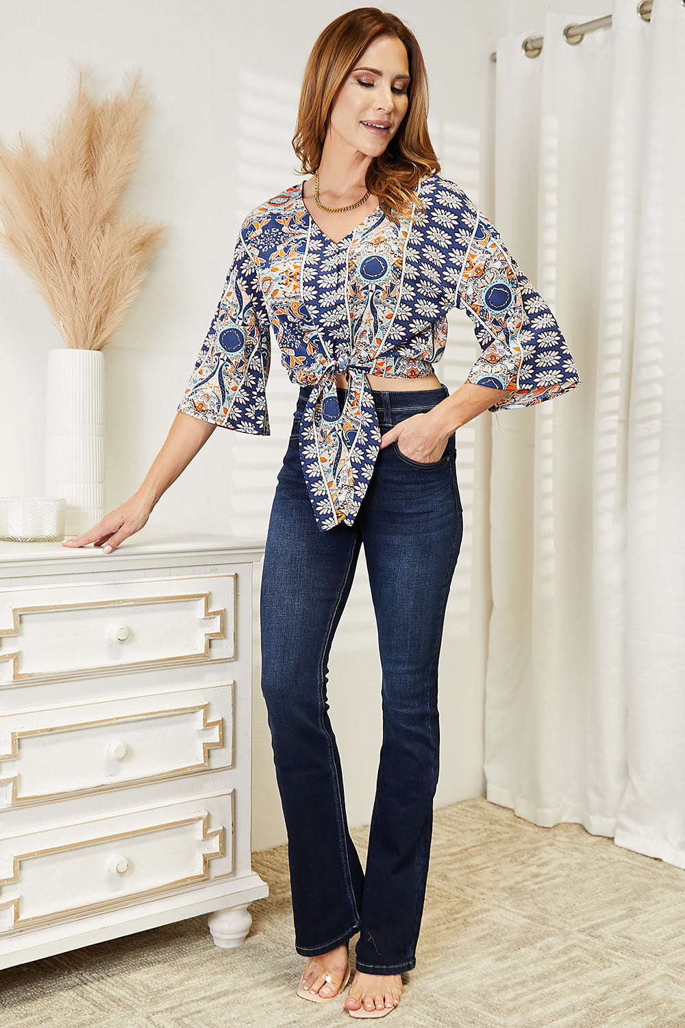 Woman wearing tie hem V-neck three-quarter sleeve blouse with patterned design, paired with dark jeans in stylish room setting.