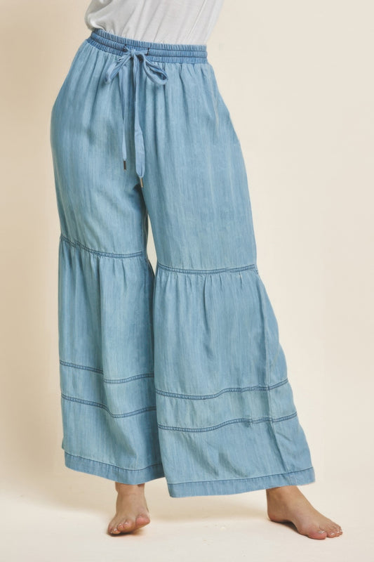 Woman wearing light blue denim wide-leg pants with tiered ruffles and drawstring waist, paired with white top, standing barefoot against neutral ba...