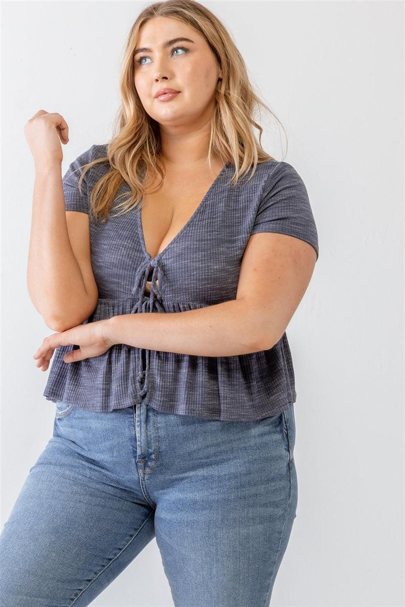 Plus size model wearing denim blue blouse with ribbed short sleeves, U-neckline, and flare hem with tie details, styled with jeans.