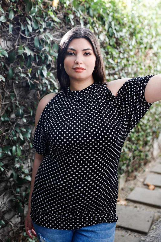 Plus size woman wearing a black polka dot open shoulder blouse with cuffed sleeves and halter neckline, posing outdoors.