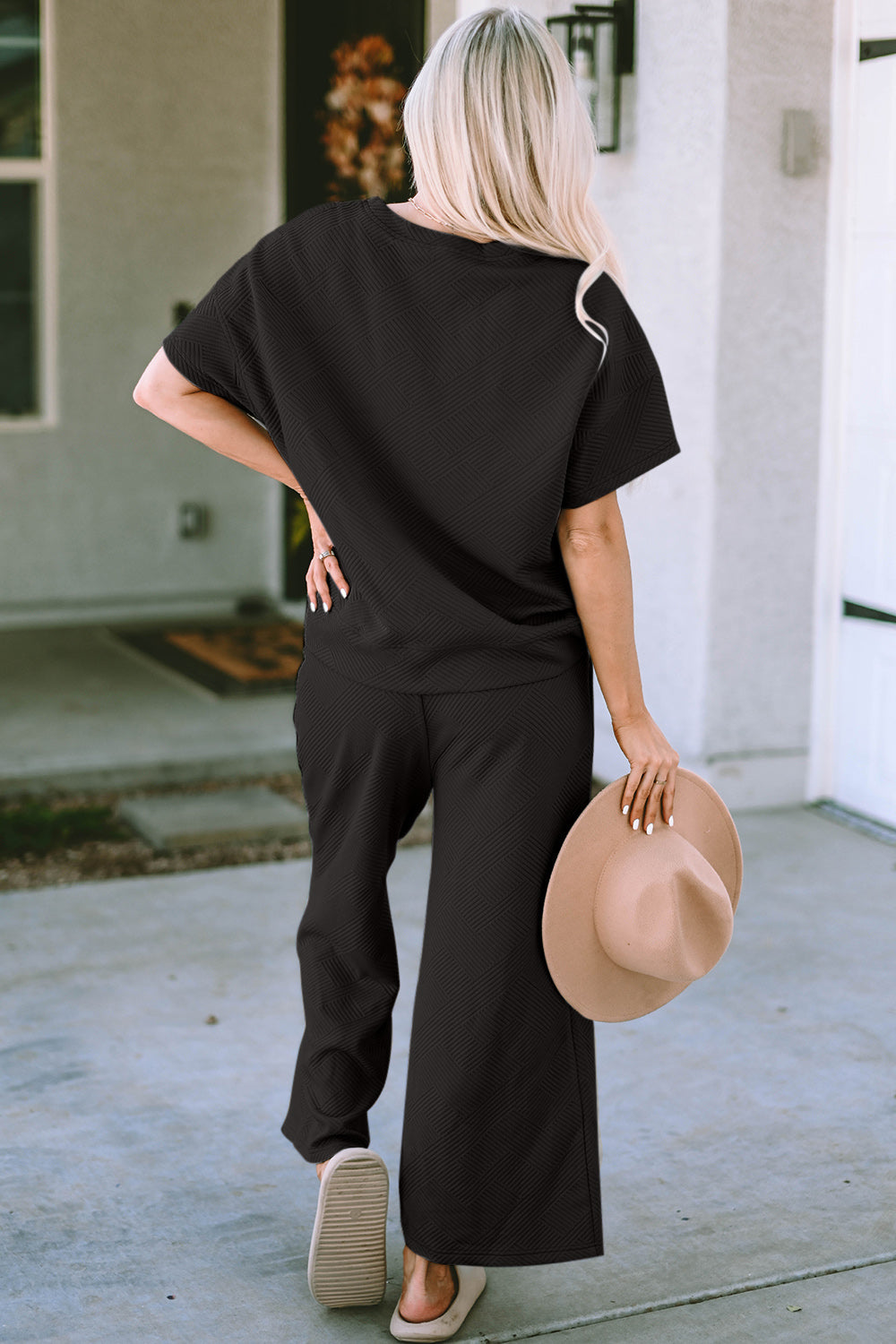 Woman wearing black short sleeve top and pants set, holding a hat, showcasing the stylish two-piece with slightly stretchy fabric.