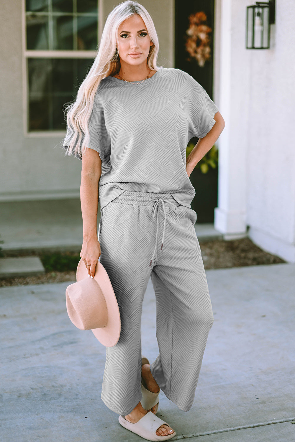 Woman in gray textured short sleeve top and pants set with drawstring, holding a hat, standing outdoors.