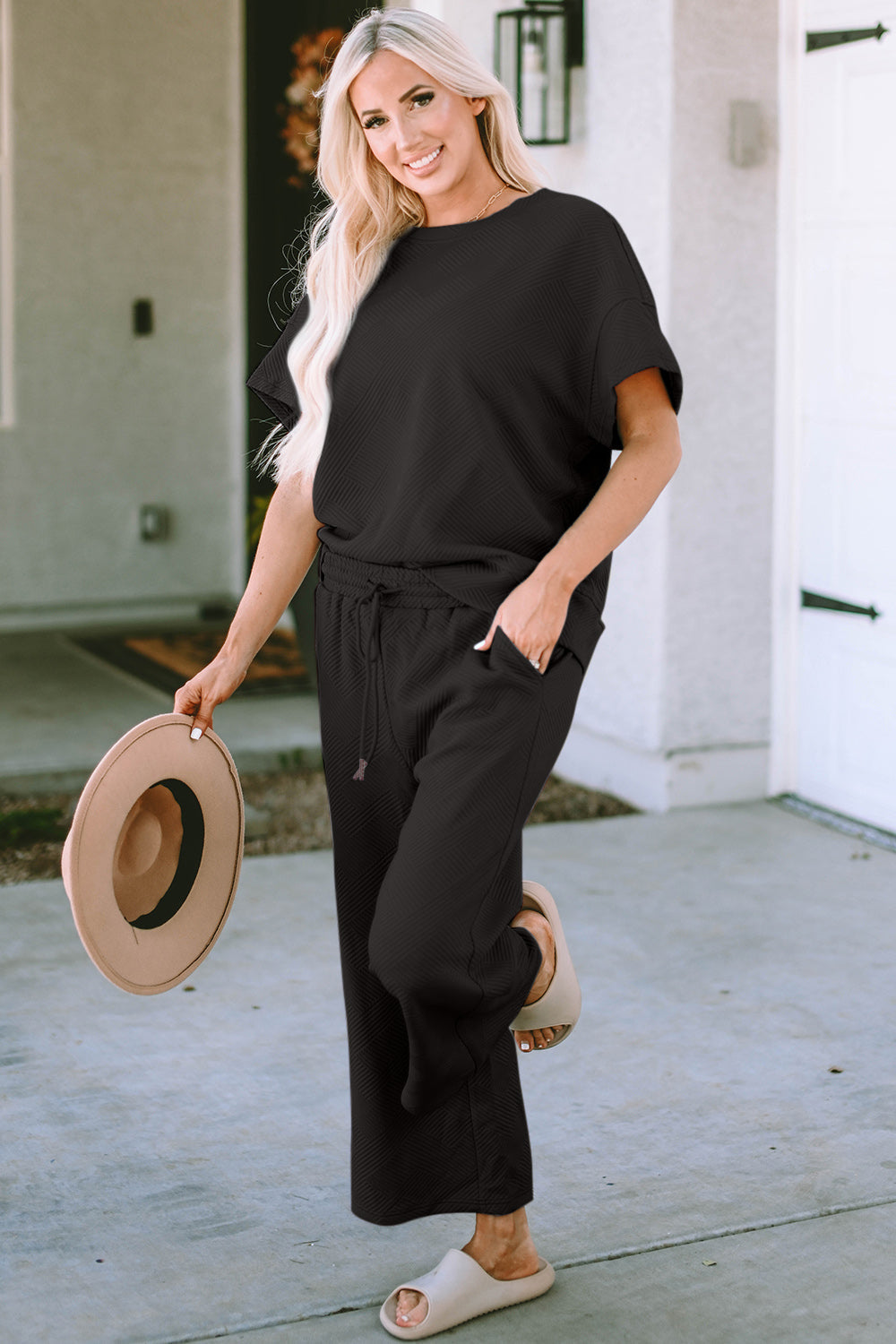 Woman wearing a Texture Short Sleeve Top and Pants Set in black with a drawstring, holding a hat. Slightly stretchy polyester-spandex blend.