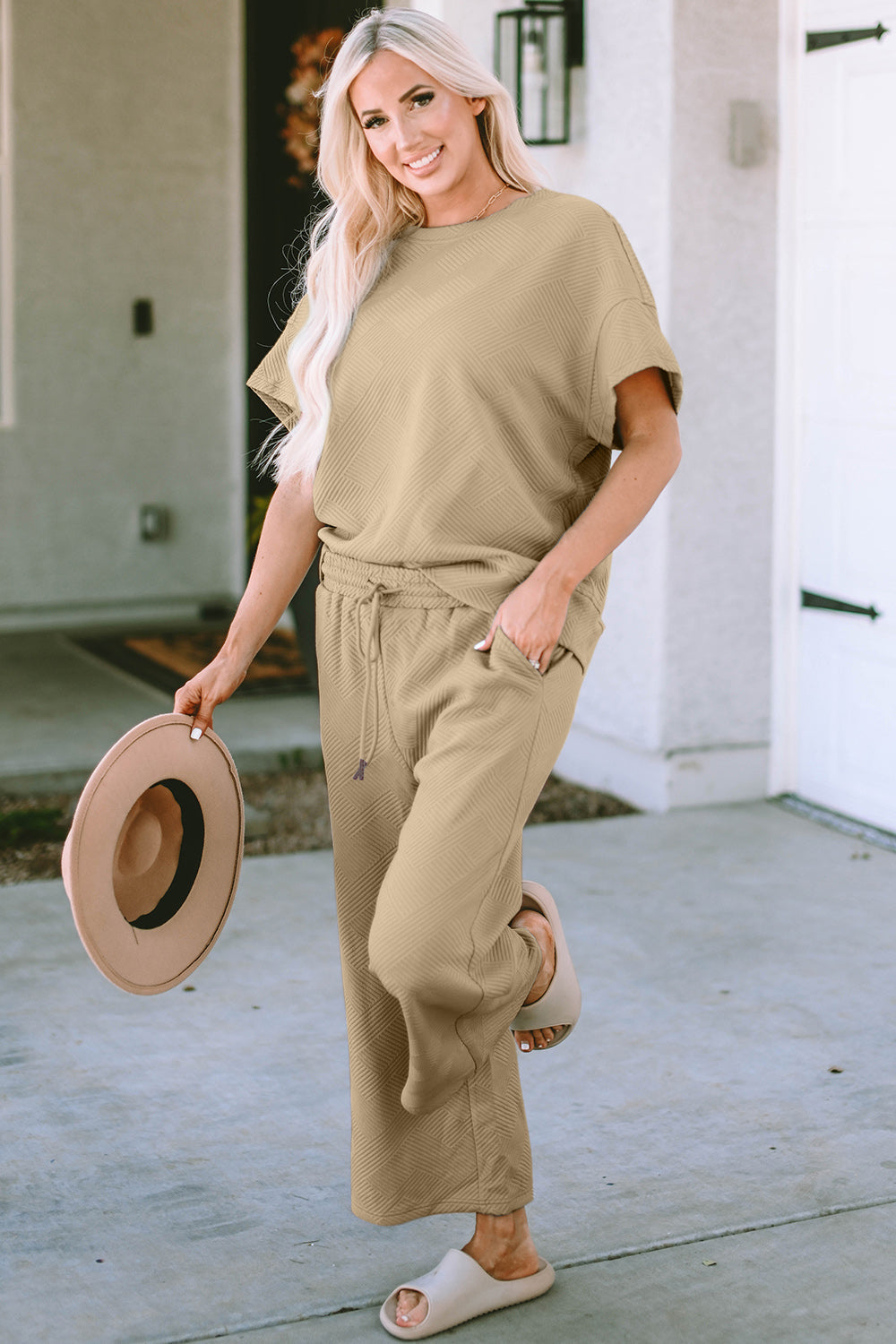 Woman wearing beige texture short sleeve top and pants set with drawstring, holding a hat, styled for casual outdoor look.