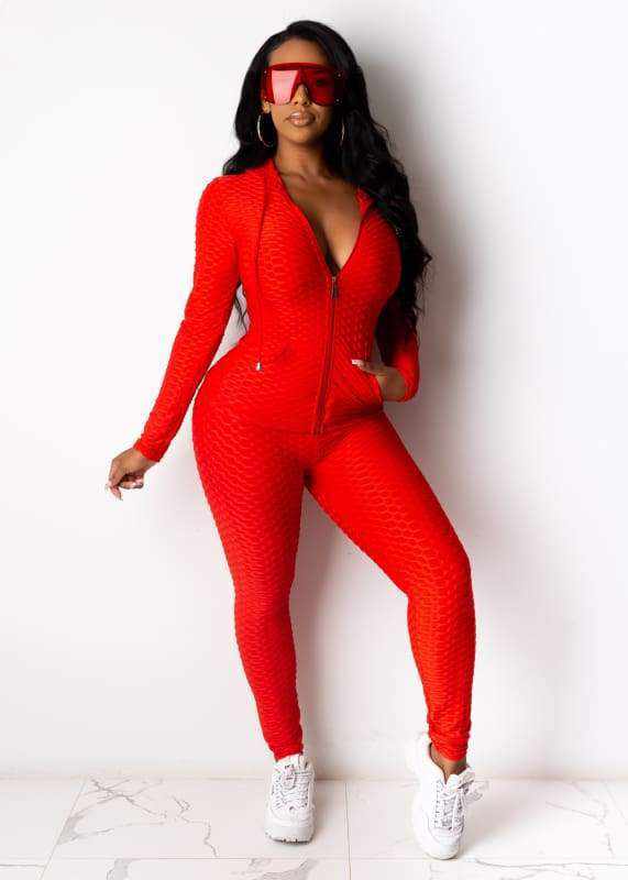 Woman wearing red textured jumpsuit and sunglasses with white sneakers, posing against a white background.