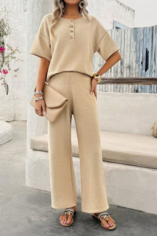 Woman in beige knit two-piece set with button-front top and wide-leg pants, holding cream handbag, wearing sandals, standing on stone steps in...