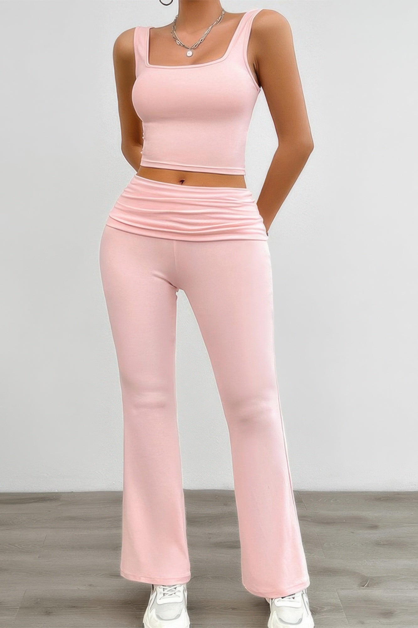 Woman wearing pink two-piece matching set with cropped tank top and high-waisted flared pants, white sneakers, and silver chain necklace, standing...