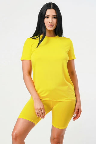 Woman with long dark hair wearing a bright yellow short-sleeve t-shirt and matching yellow bike shorts, standing against a light gray background.