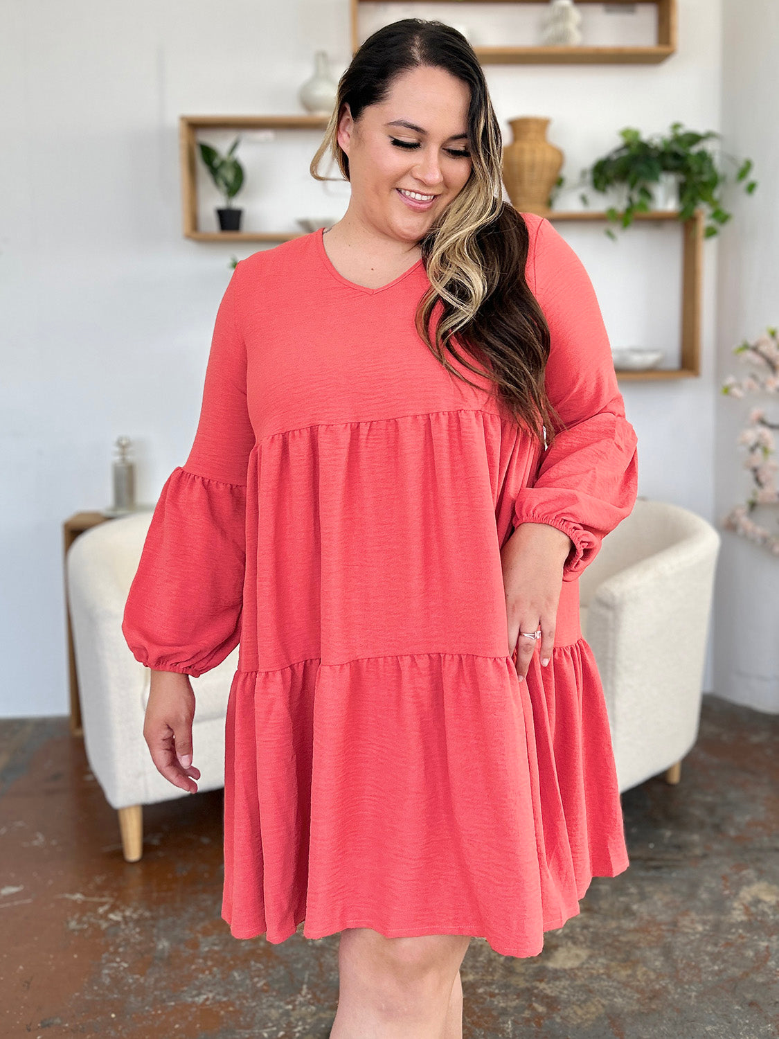 Woman wearing coral V-neck balloon sleeve tiered dress with pockets, showcasing its ruffled, pocketed design in a stylish room setting.