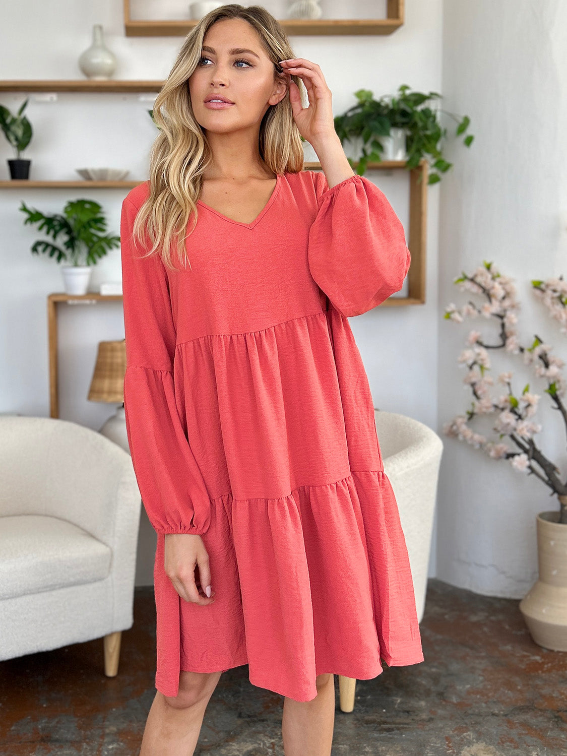 Woman wearing a V-neck balloon sleeve tiered dress with pockets in coral color, standing in a stylish room with plants.