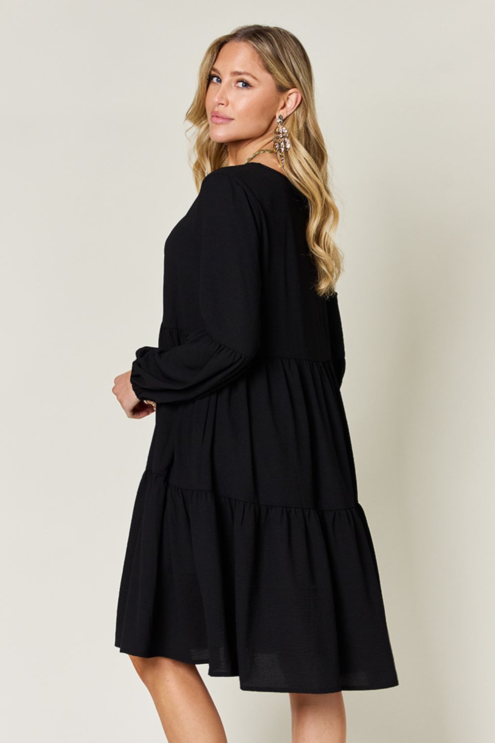 Woman wearing a black V-neck balloon sleeve tiered dress with pockets and ruffles, made from high stretch yarn, viewed from the side.