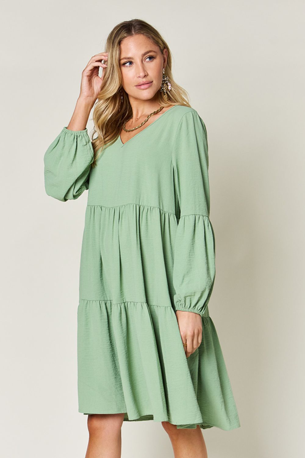 Woman wearing a green V-neck balloon sleeve tiered dress with pockets, made of high stretch yarn, featuring an opaque, ruffled fit.