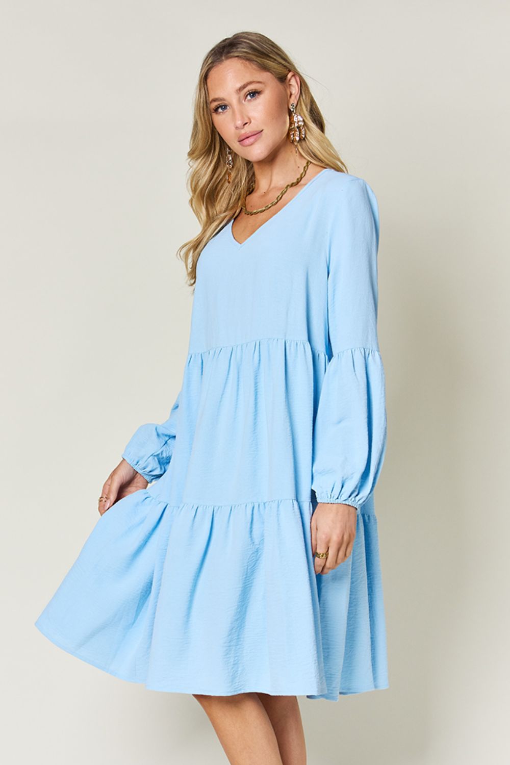 Woman wearing a blue V-neck balloon sleeve tiered dress with pockets, ruffled details, made from 100% high stretch yarn.