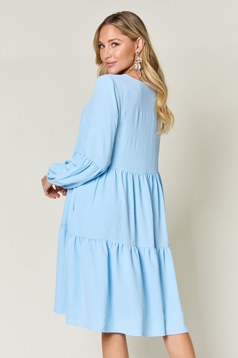 Woman wearing a light blue V-neck tiered dress with balloon sleeves and pockets, showing the back view.