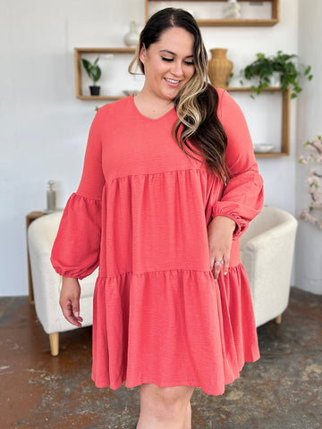 Woman wearing coral V-neck balloon sleeve tiered dress with pockets, showcasing its ruffled, pocketed design in a stylish room setting.