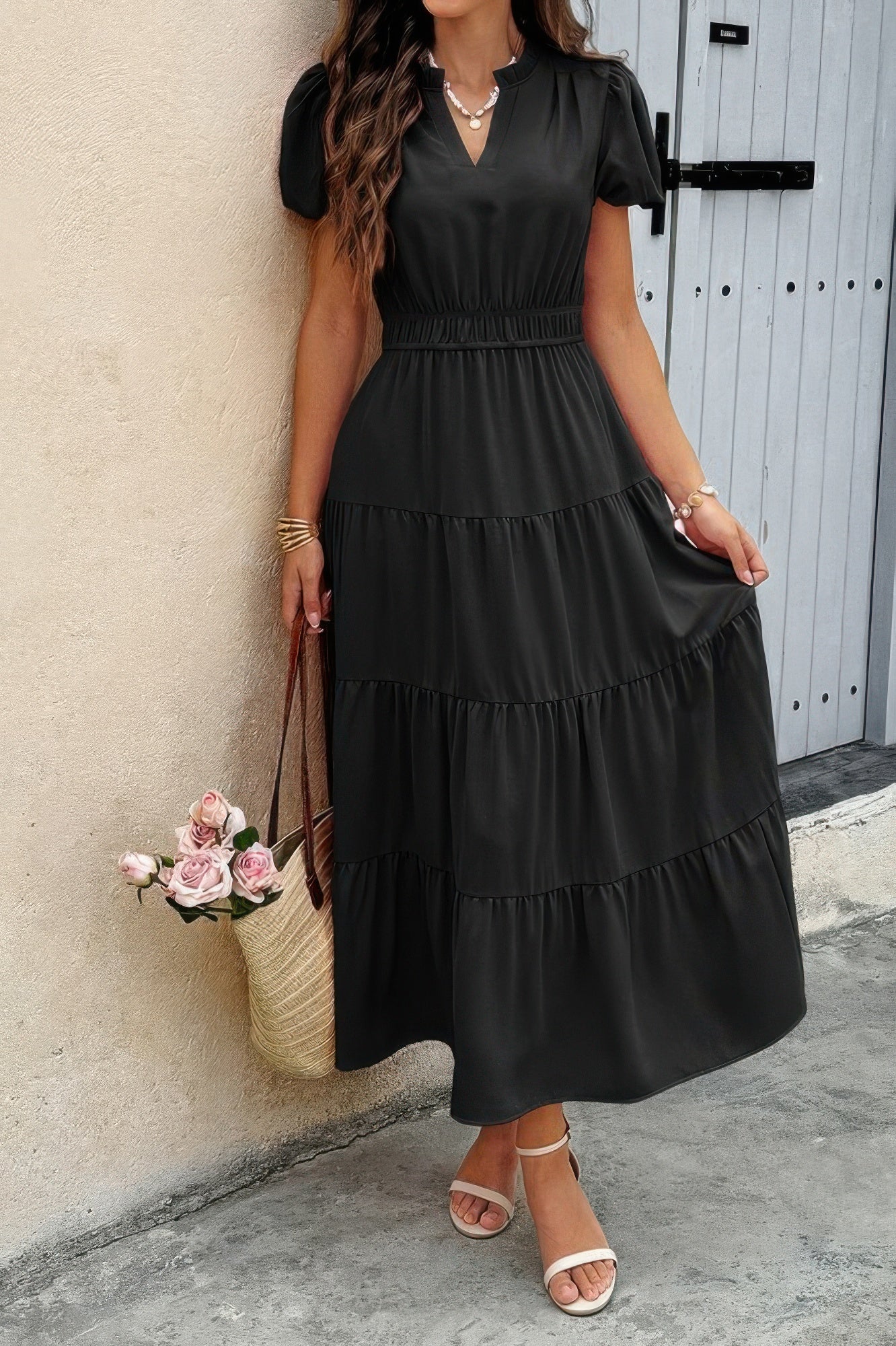 Woman in black tiered maxi dress with short sleeves and V-neck, wearing white sandals and gold jewelry, holding woven bag with pink roses, standing...