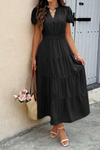 Woman in black tiered maxi dress with short sleeves and V-neck, wearing white sandals and gold jewelry, holding woven bag with pink roses, standing...