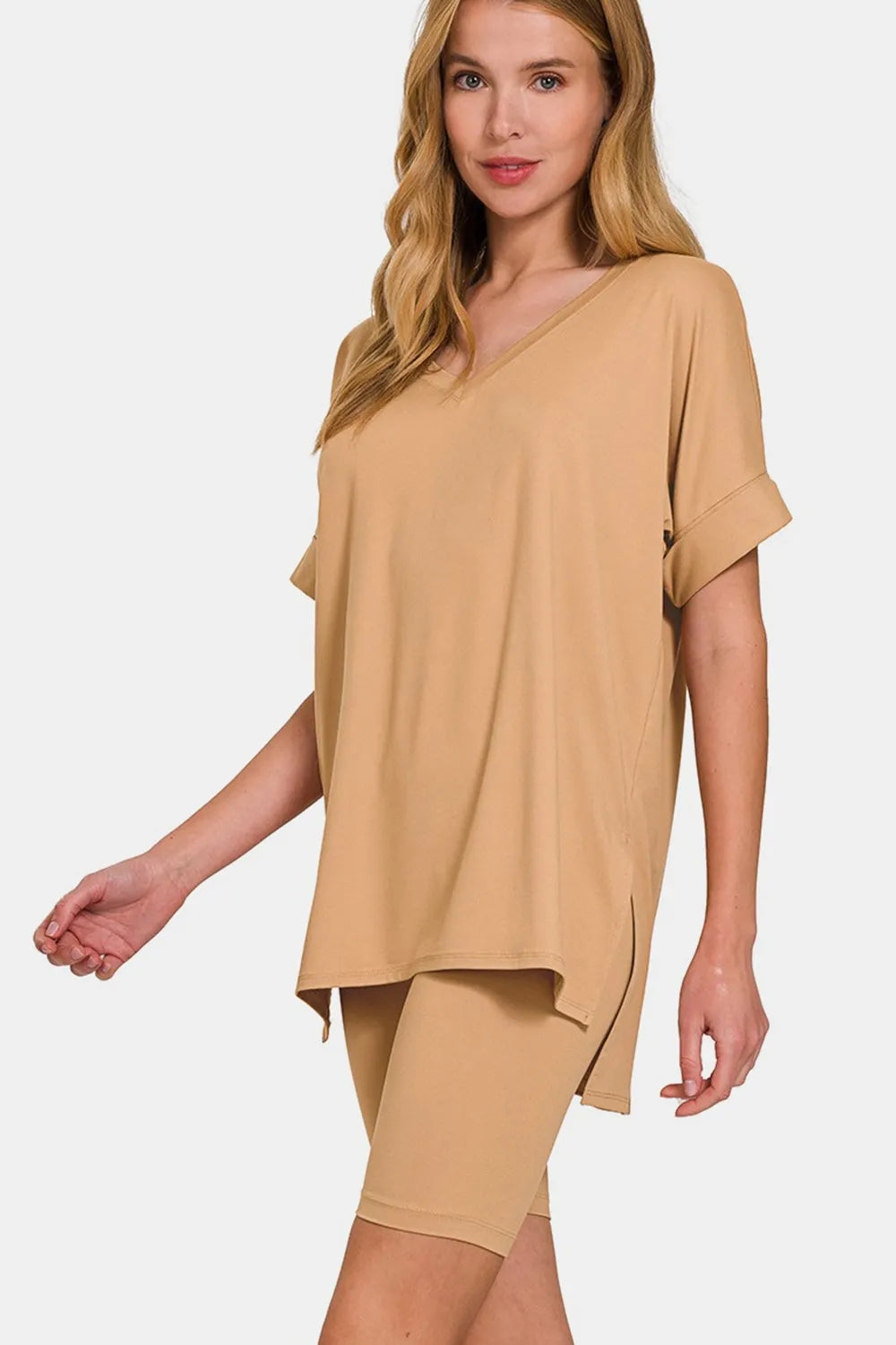 Woman with blonde hair wearing a tan/beige oversized short-sleeve top and matching shorts, standing against a white background with one hand on hip.