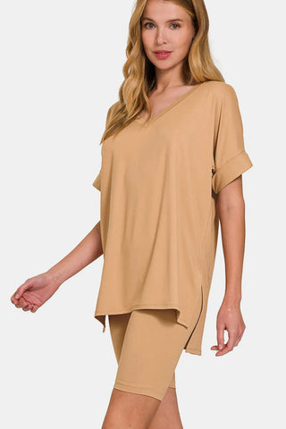 Woman with blonde hair wearing a tan/beige oversized short-sleeve top and matching shorts, standing against a white background with one hand on hip.