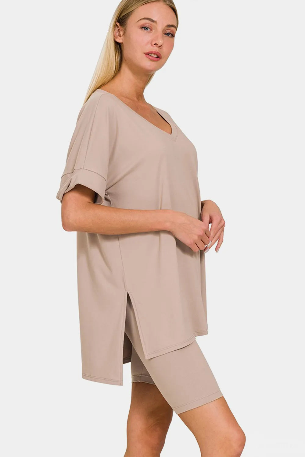 Woman with long blonde hair wearing a beige two-piece loungewear set consisting of an oversized V-neck top and shorts, posed against a white...