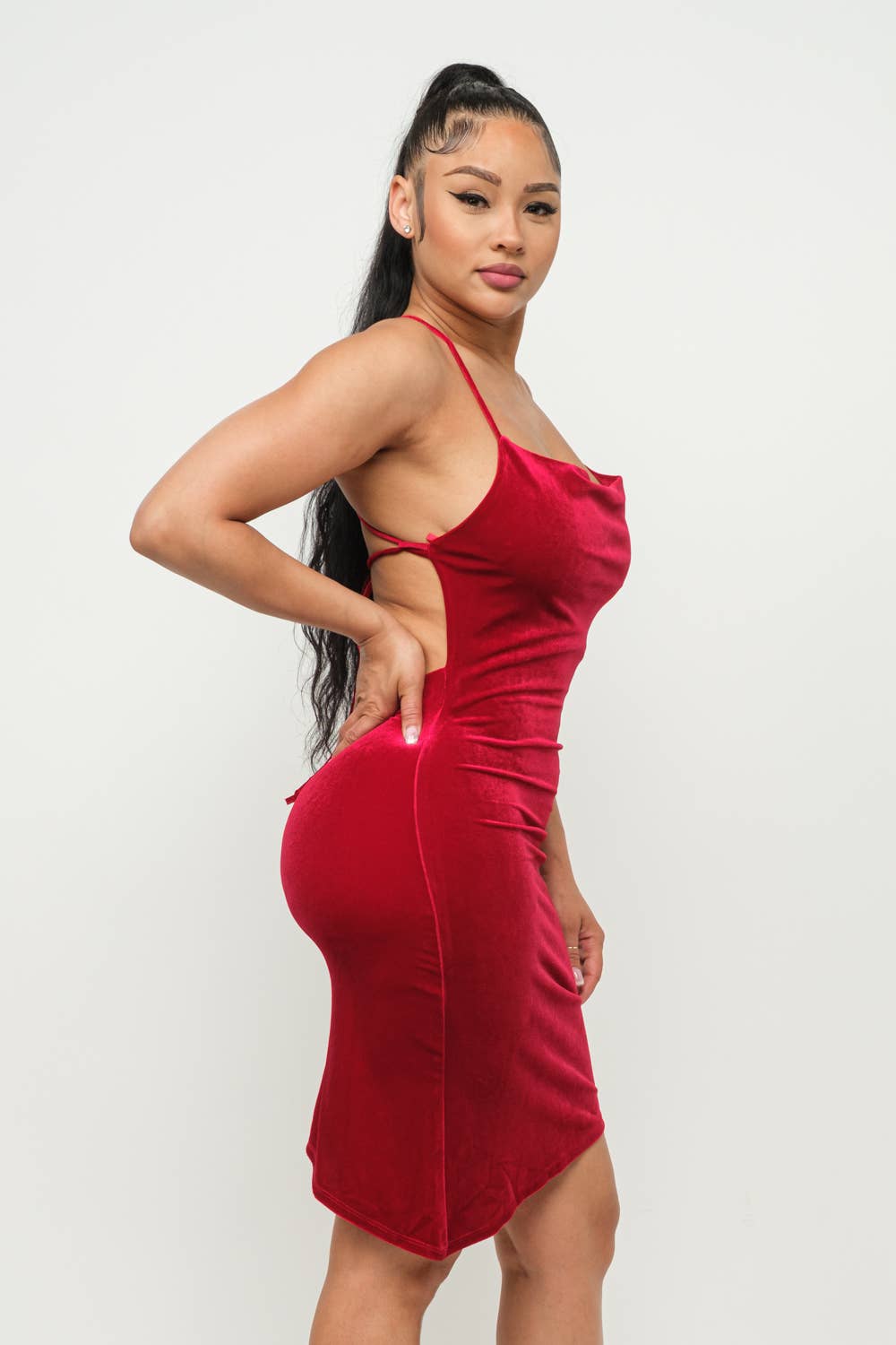 Woman wears red velvet spaghetti strap cross back bodycon dress with asymmetrical hem, showcasing side tie detail and cowl neck.