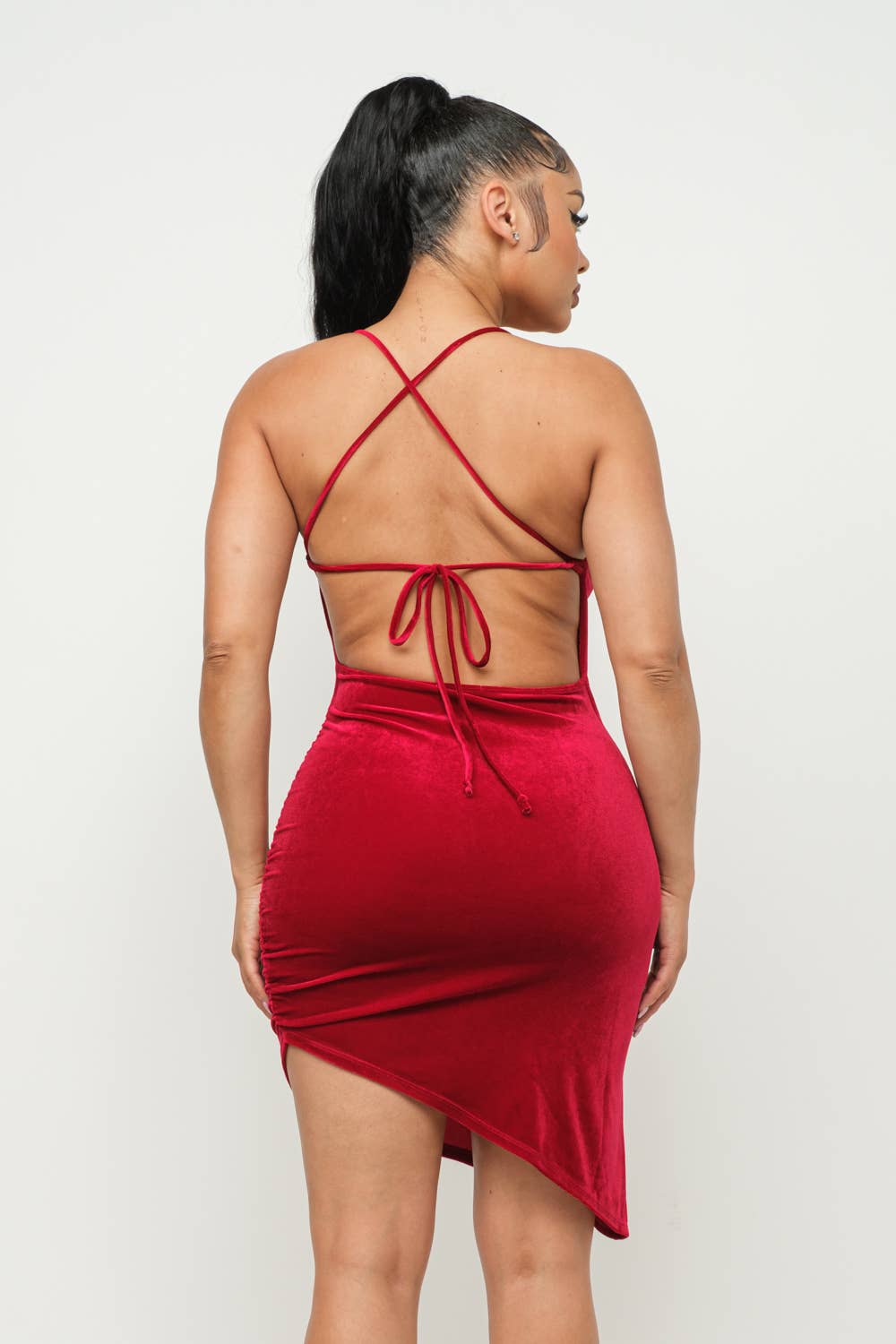 Woman wearing red velvet cross back tie side asymmetrical hem dress, showing back details and bodycon fit.