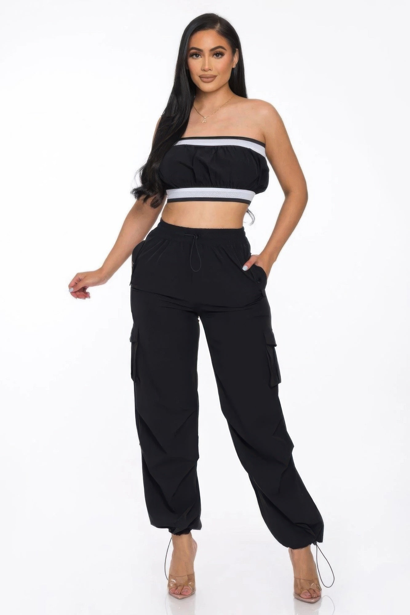 Woman in black strapless crop top with white trim and matching black cargo pants, standing against white background. She has long dark hair and is ...