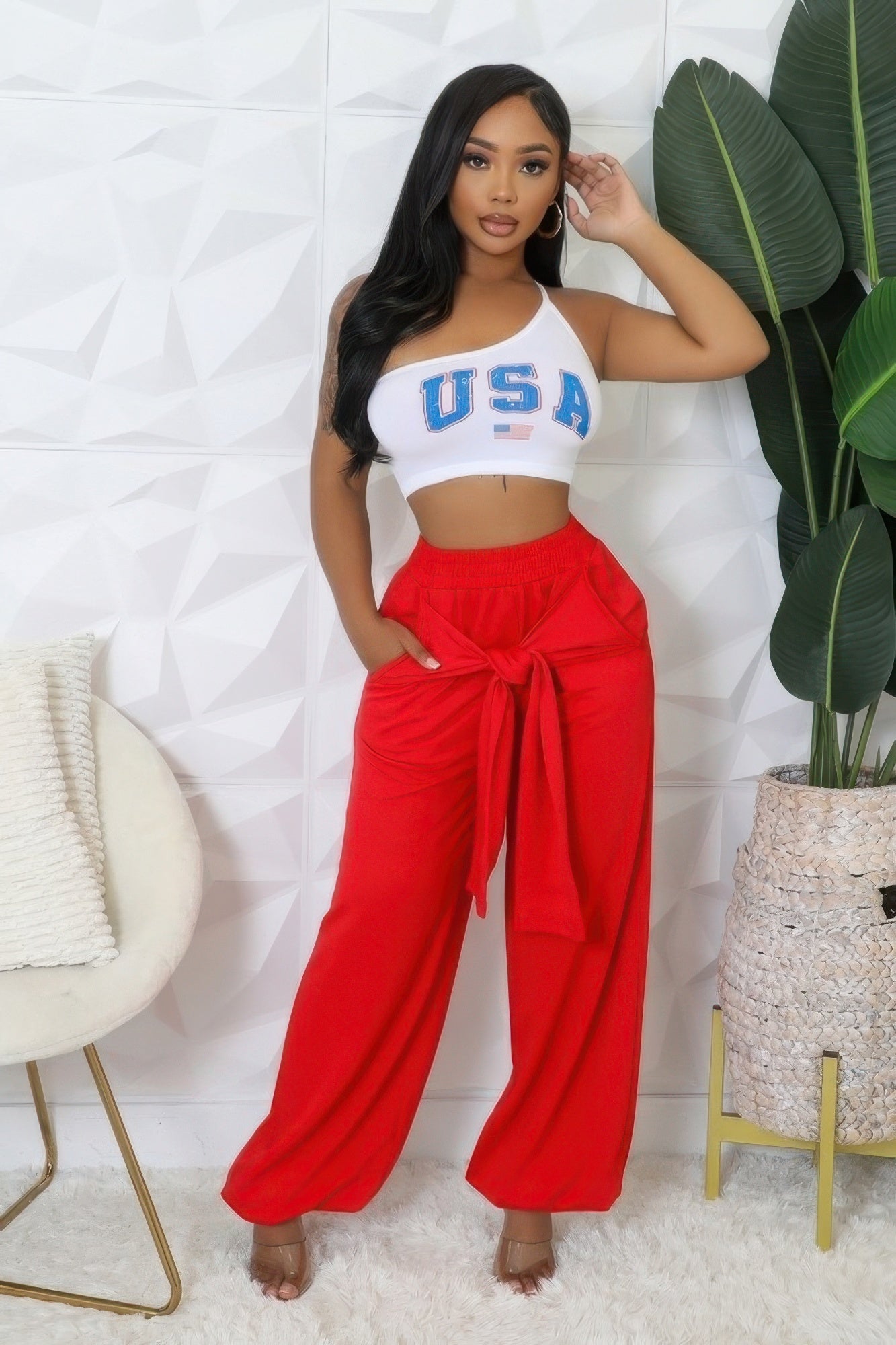 Woman wearing red high-waisted pants with a white crop top, posing in a modern room with plants and stylish decor.