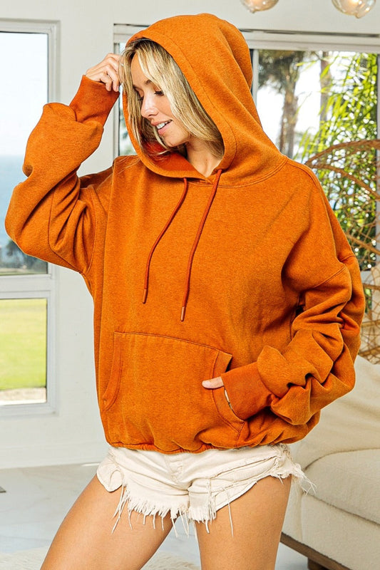 Woman wearing an orange ruched long sleeve washed fleece hoodie, standing indoors with a relaxed look.