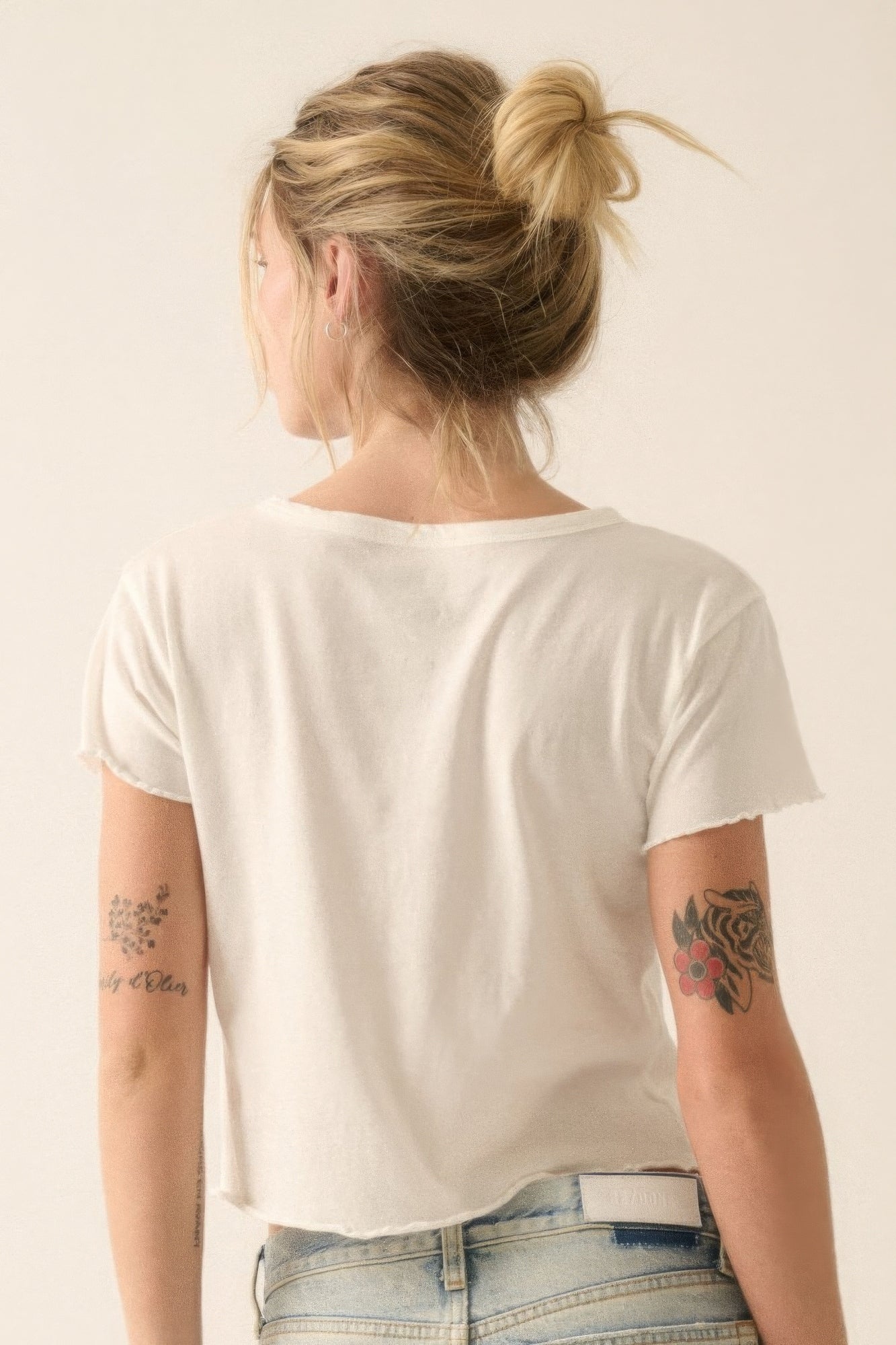 Woman wearing cropped vintage wash graphic tee in ivory, with tattoos visible, styled with light denim jeans and casual bun hairstyle.