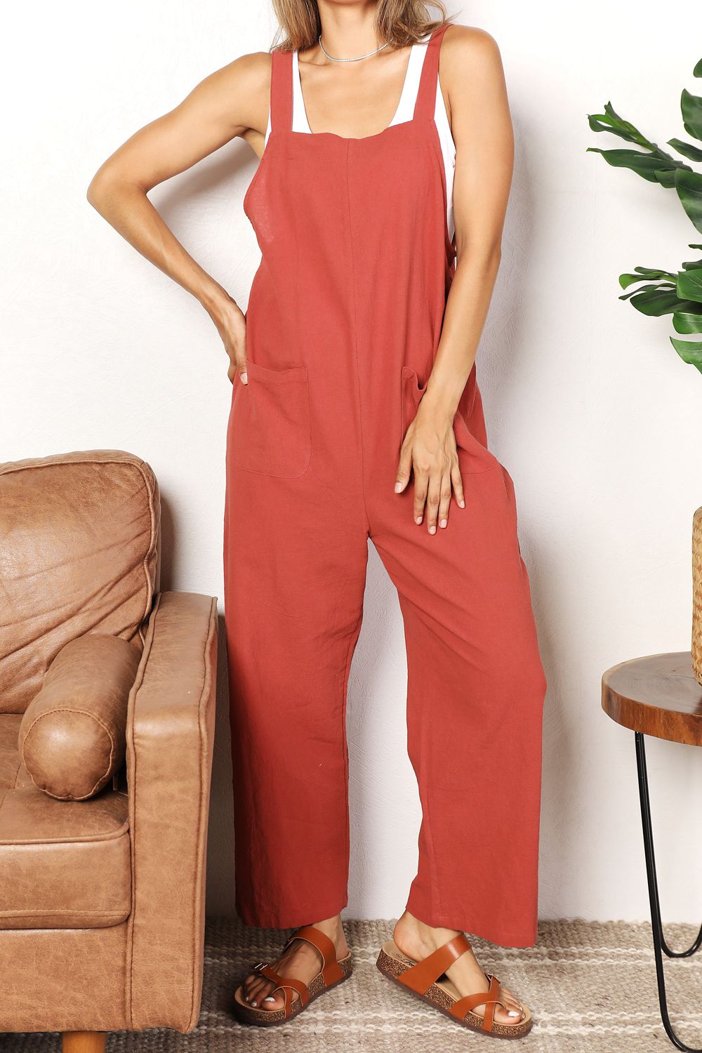 Woman wearing chic wide leg overalls with front pockets, styled with sandals, standing in a cozy living room setting.