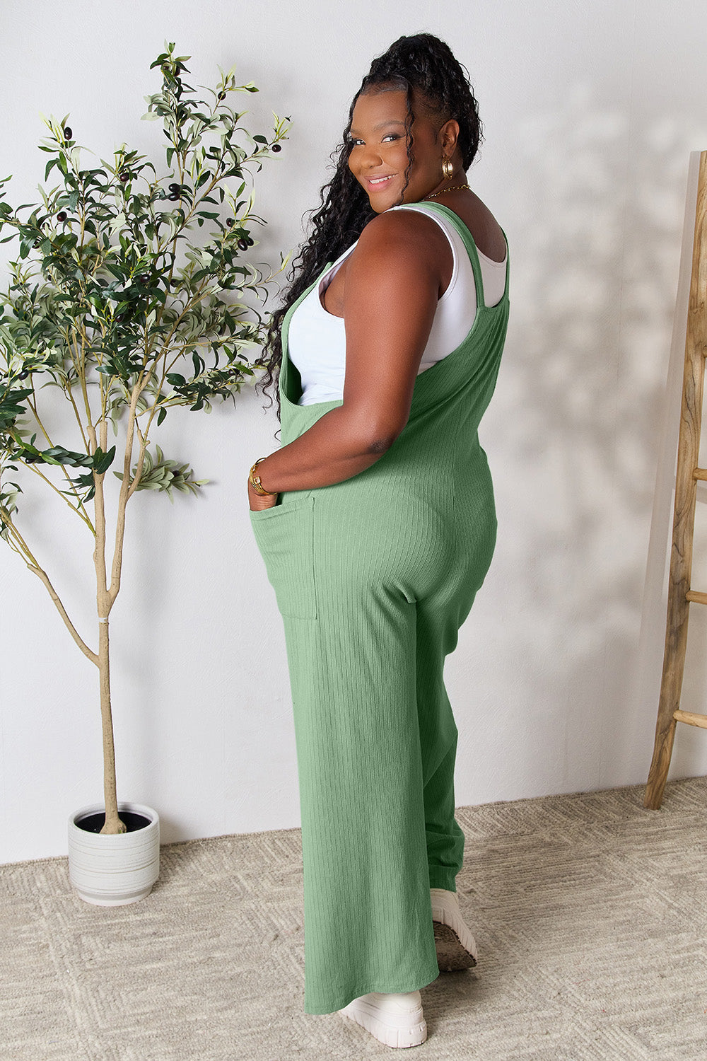 Woman wearing a green wide strap overall with pockets, made of polyester, rayon, and spandex, standing near a potted plant.